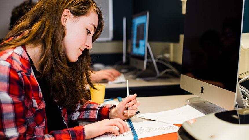 A student works with paper and pencil in front of a computer